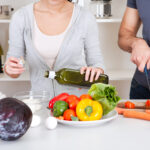 Couple cooking together with a variety of fresh bell peppers, cabbage, and other vegetables, drizzling olive oil.