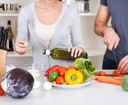 Couple cooking together with a variety of fresh bell peppers, cabbage, and other vegetables, drizzling olive oil.